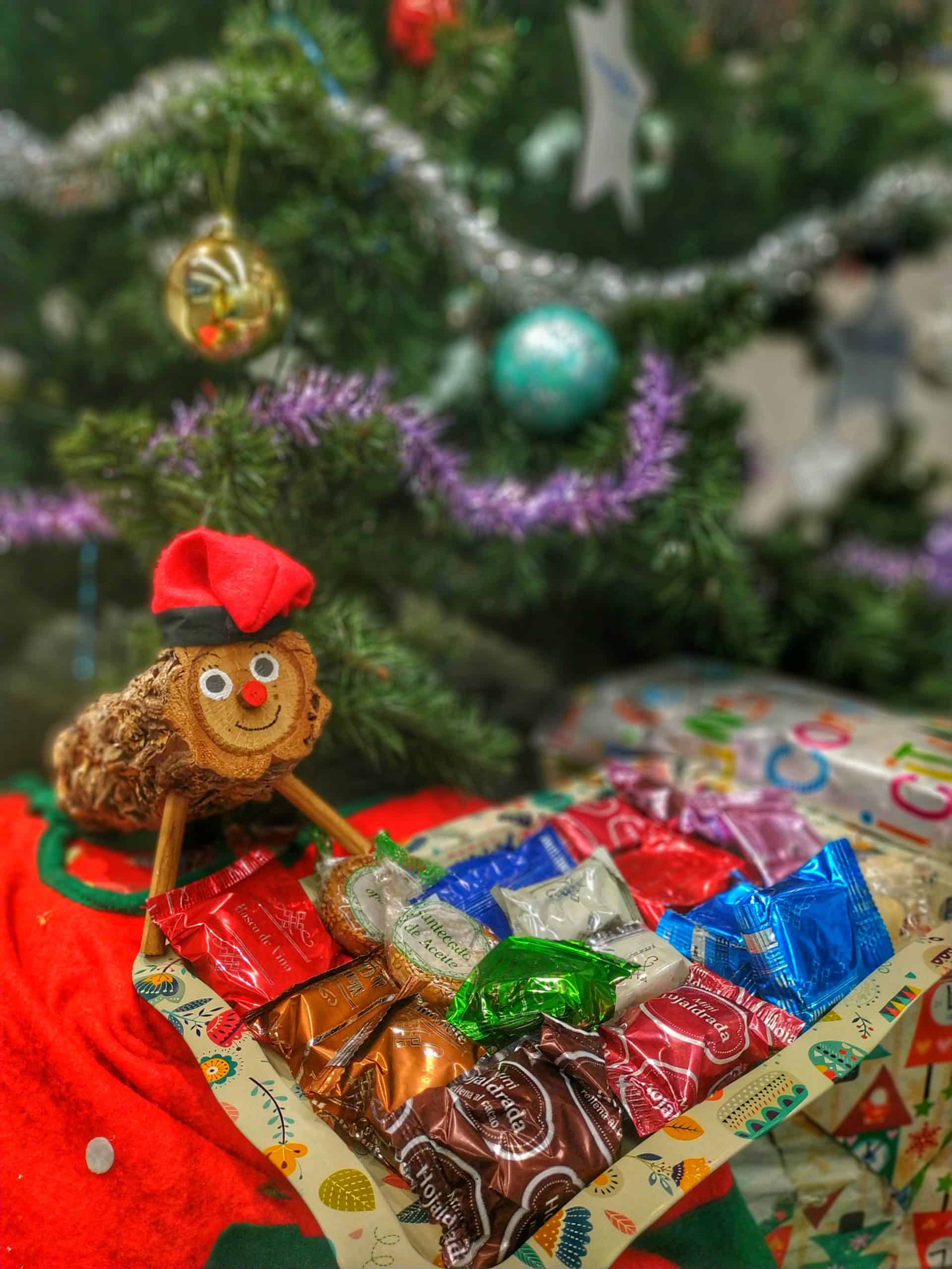 Tió de Nadal rodeado de dulces bajo un árbol de Navidad decorado.