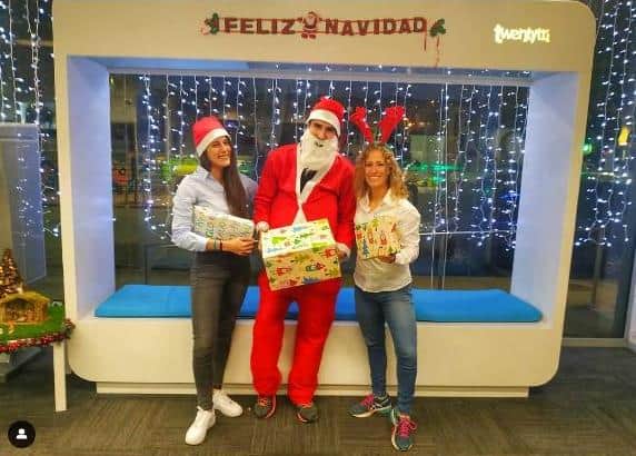 Papá Noel y su equipo para Navidad en TwentyTú Hostel sosteniendo regalos en un espacio decorado con luces y un cartel de “Feliz Navidad”.