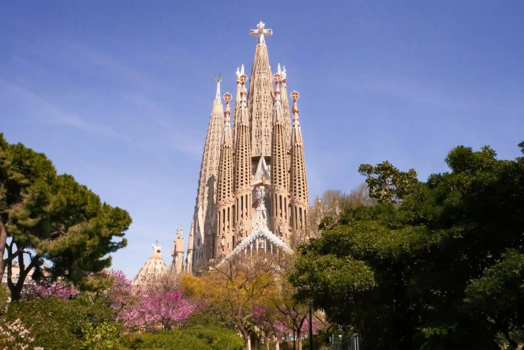 Vista exterior de la Basílica de la Sagrada Familia rodeada de árboles y flores bajo un cielo despejado en Barcelona.