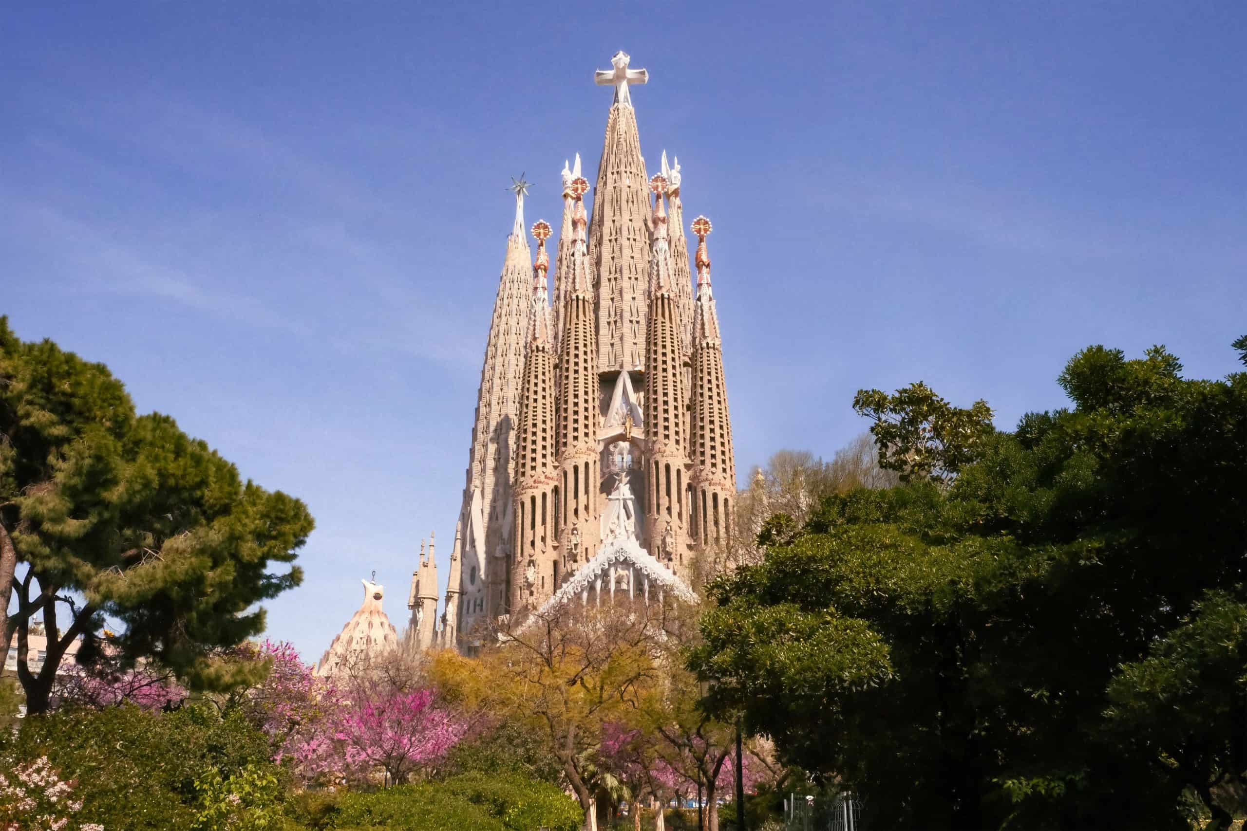 Vista exterior de la Basílica de la Sagrada Familia rodeada de árboles y flores bajo un cielo despejado en Barcelona.